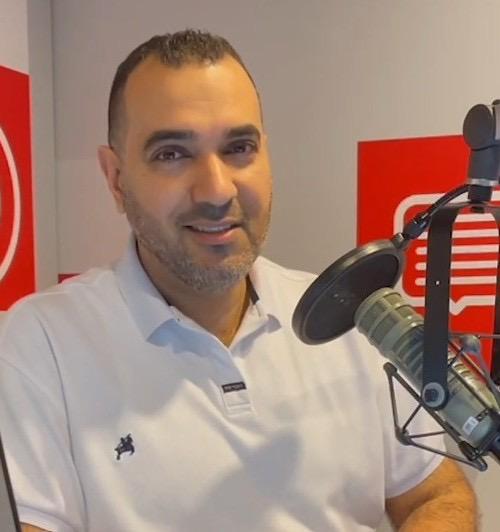 رجل يجلس أمام ميكروفون في استوديو إذاعي، مبتسم، يرتدي قميصاً أبيض.