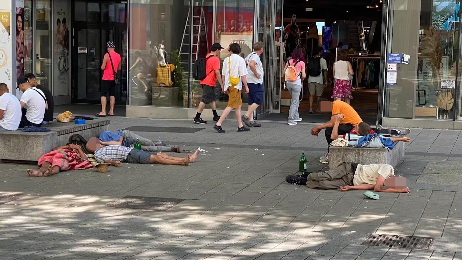 مجموعة من الأشخاص مستلقين على الأرض في منطقة حضرية، مع وجود بعض المارة في الخلفية، مما يعكس مشاكل اجتماعية في المدينة.