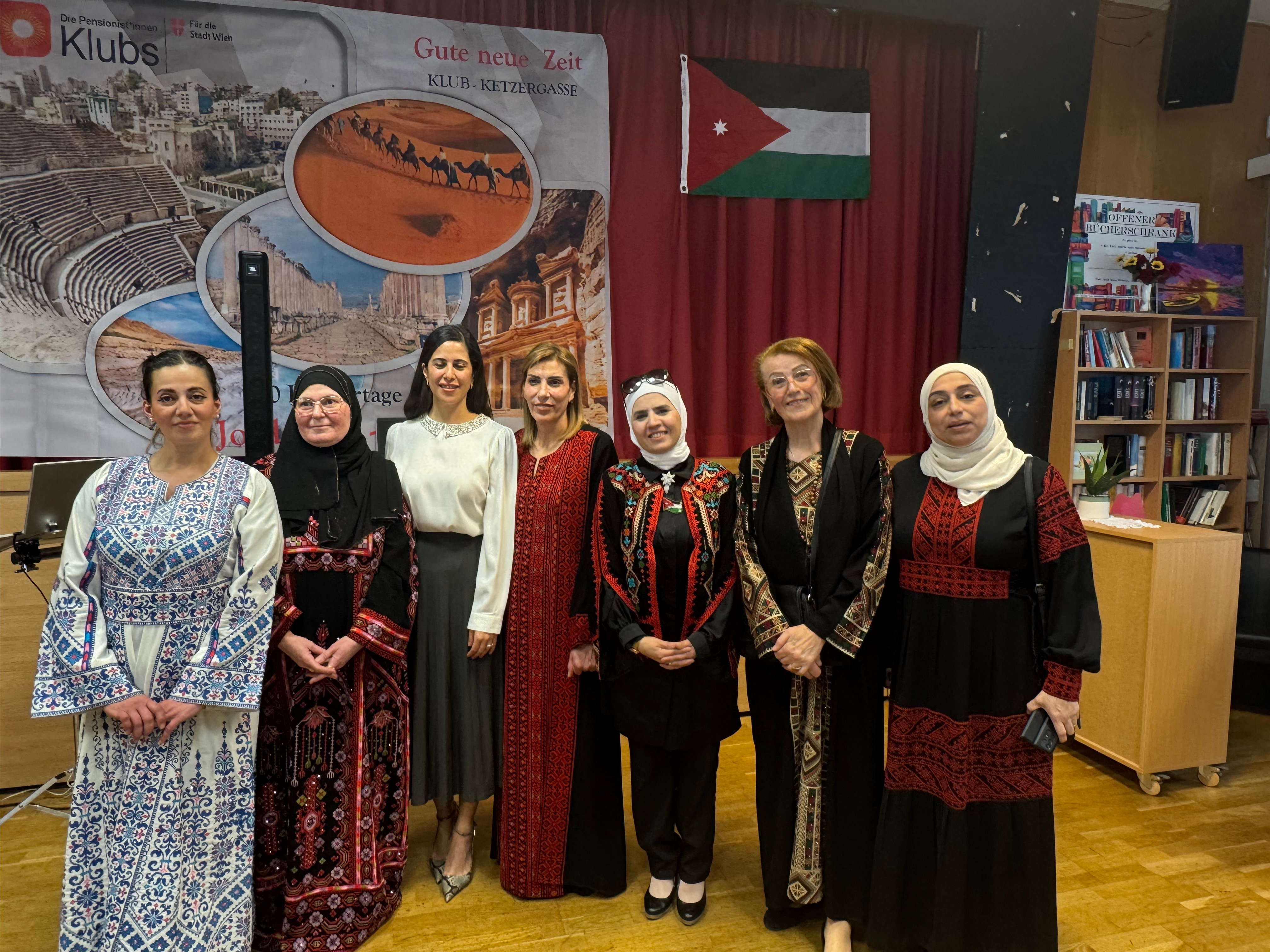 مجموعة من النساء يرتدين أزياء تقليدية متنوعة، يقفن معًا في حدث ثقافي. خلفهم علم الأردن ومشهد من معالم تاريخية على الجدار.