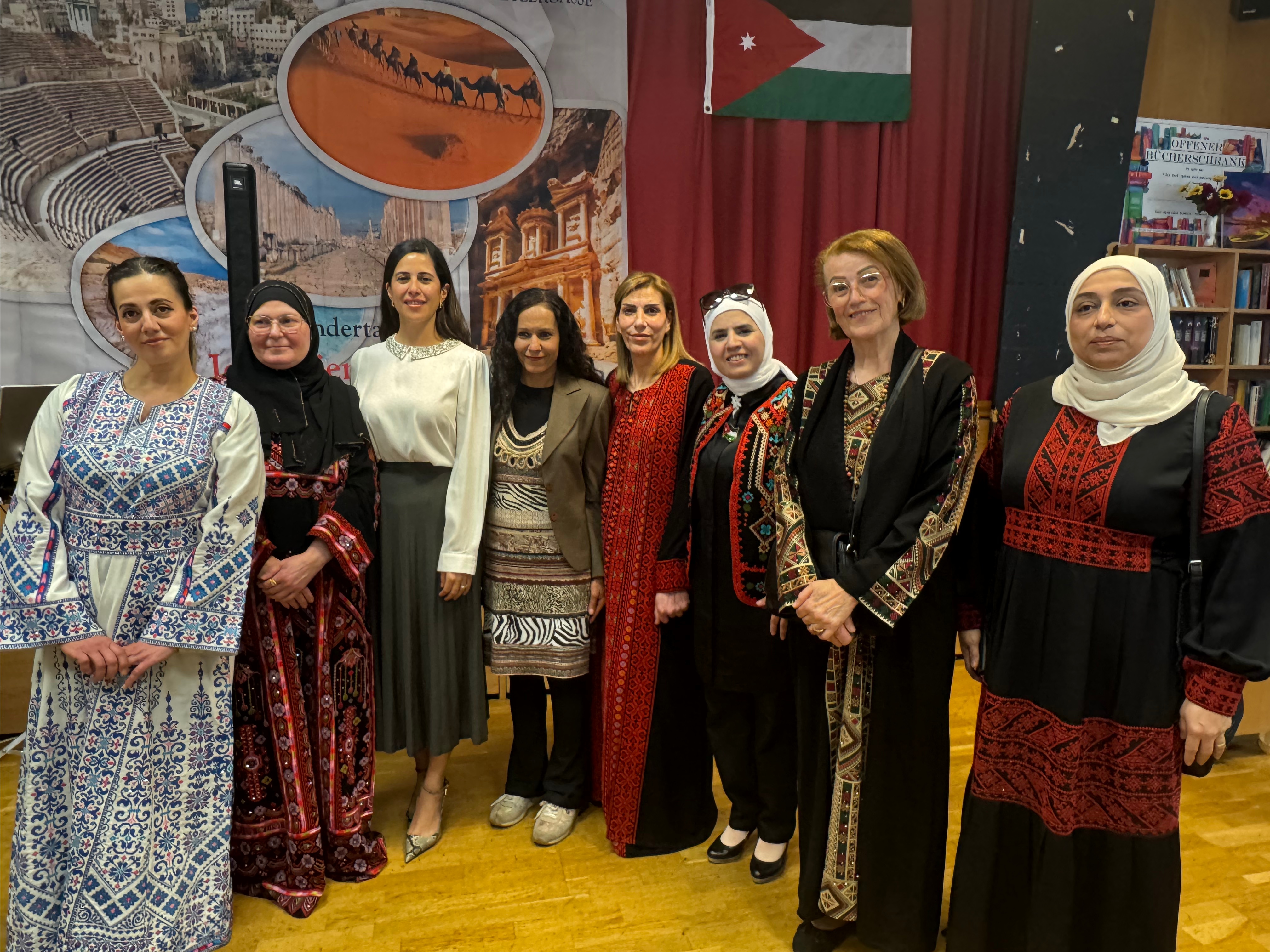 مجموعة من النساء يرتدين أزياء تقليدية متنوعة، يقفن معًا أمام خلفية تحتوي على صور لمواقع تاريخية وأعلام، ويظهرن في قاعة احتفالية.