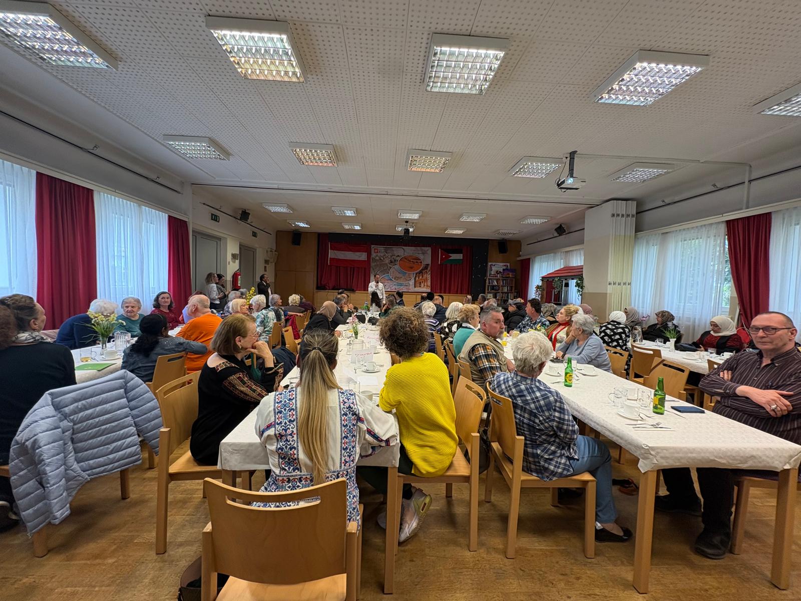 مجموعة من الأشخاص يجلسون حول طاولات في قاعة، يتناولون الطعام ويتبادلون الحديث. المكان مزين بستائر حمراء ونوافذ كبيرة تضيء الغرفة.