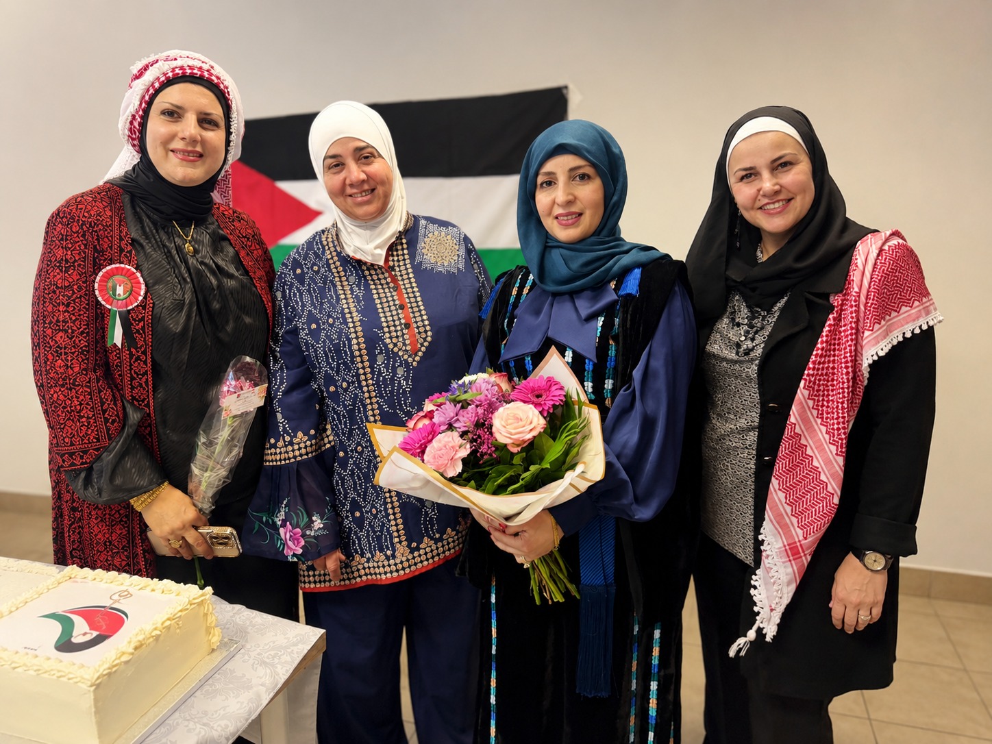 مجموعة من النساء يرتدين ملابس تقليدية ويحتفلن، مع باقة من الزهور وكعكة في المقدمة. يوجد علم فلسطين خلفهن.