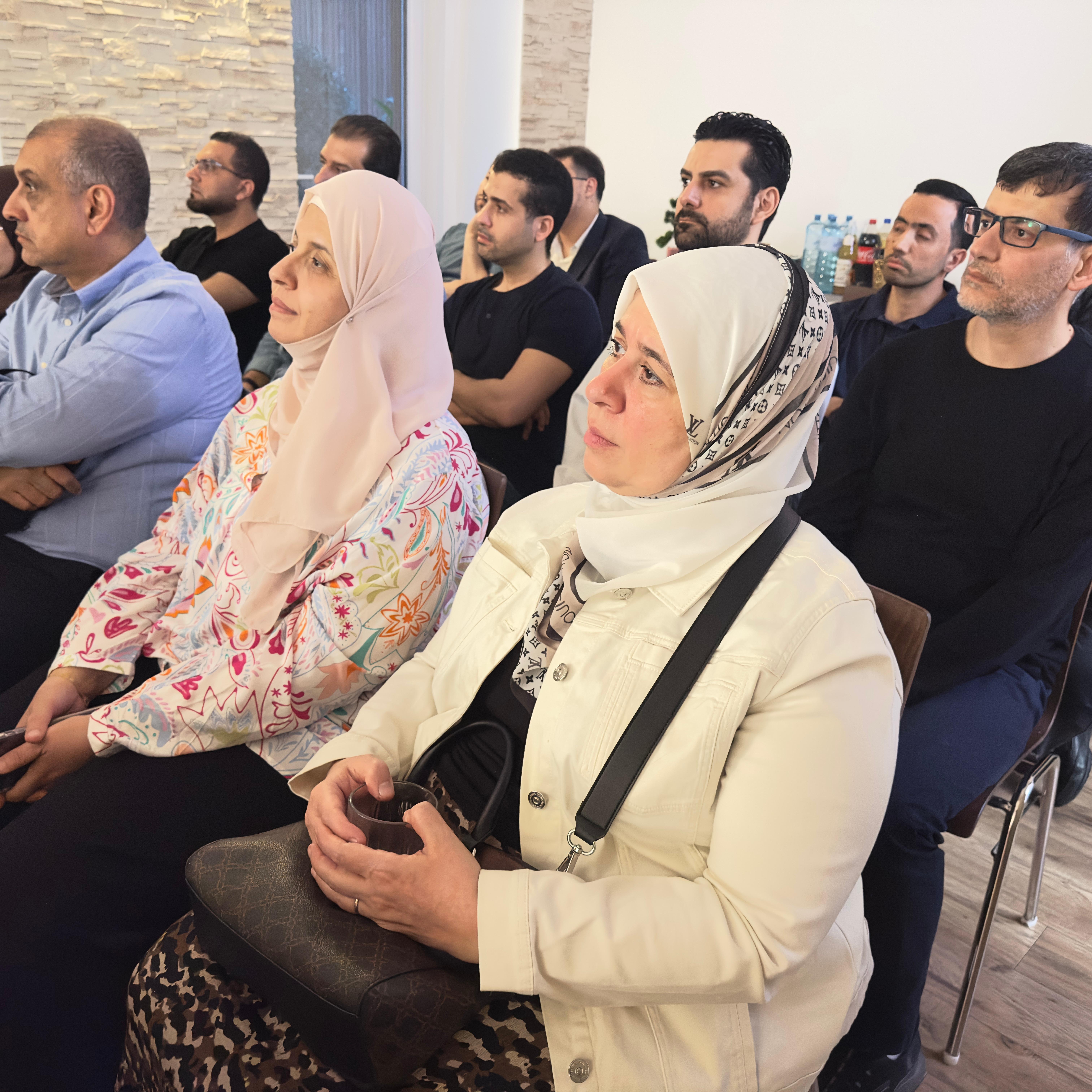 مجموعة من الأشخاص يجلسون في قاعة، يركزون على المتحدث. يظهر بعضهم بأزياء متنوعة، مع اهتمام واضح على وجوههم.