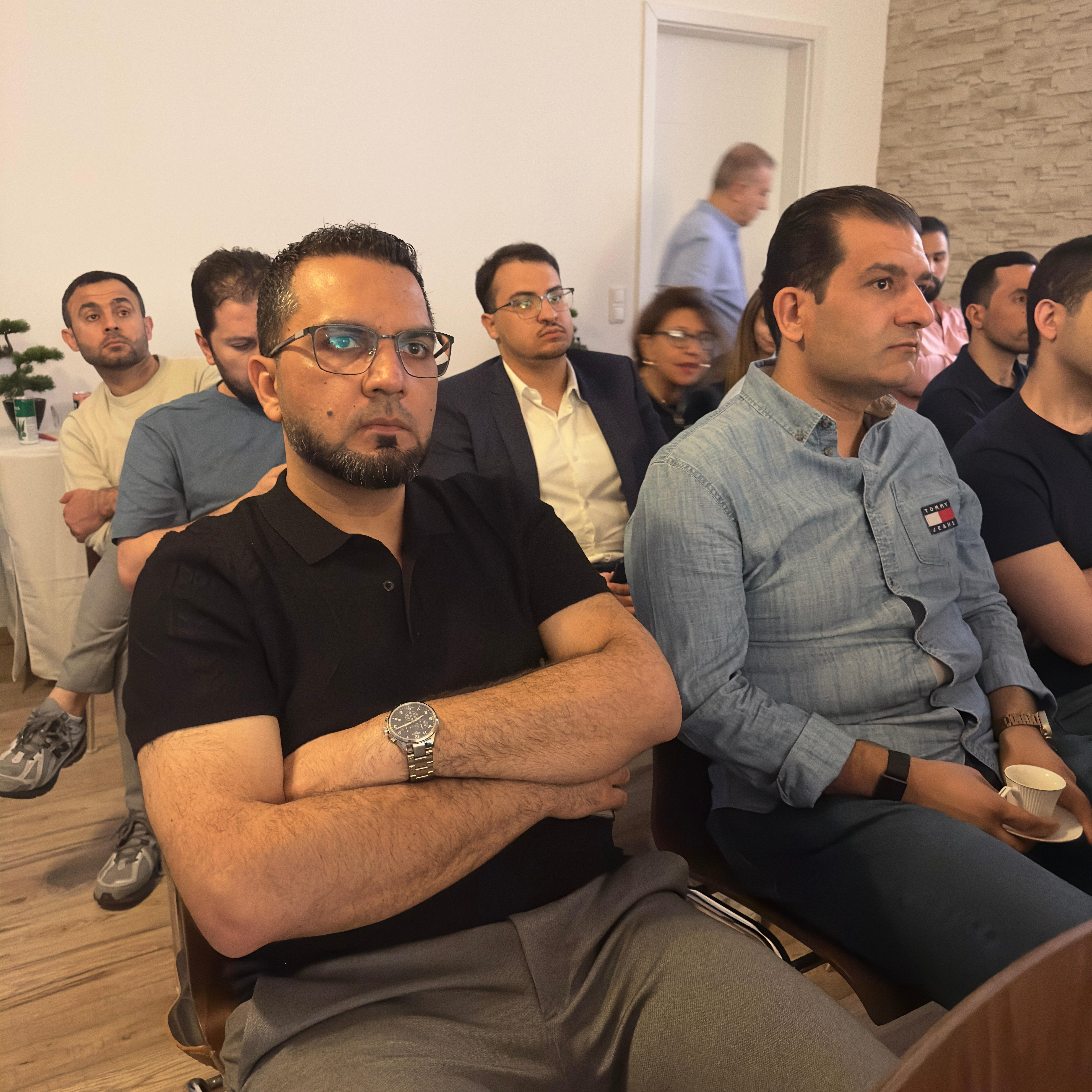 مجموعة من الرجال يجلسون في جلسة، يظهر بعضهم مهتمين بينما يبدو البعض الآخر مشغولين. رجل ذو لحية قصيرة ونظارات يجلس في المقدمة على كرسي، مع ذراعيه متقاطعتين.